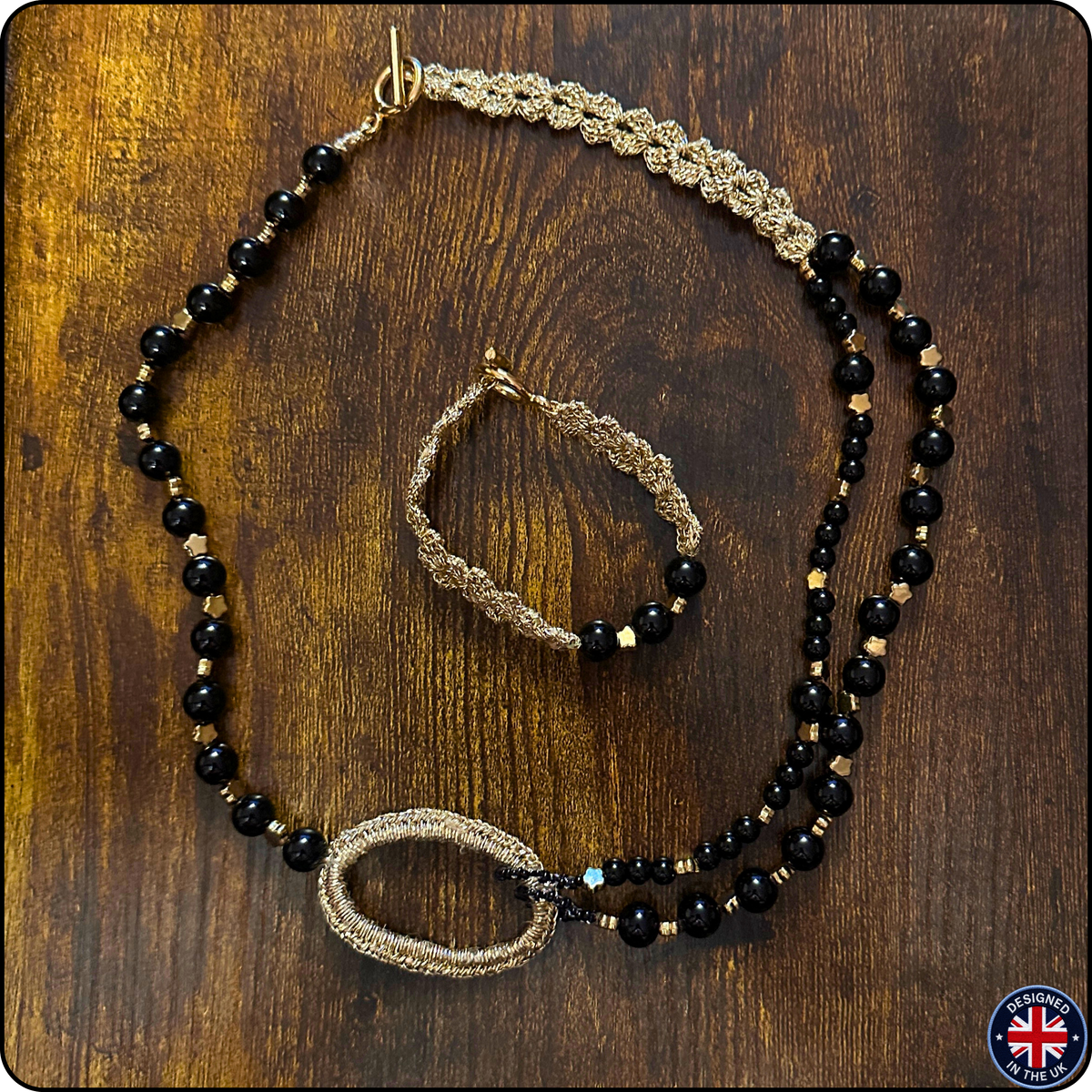 A jewelry set featuring a beaded necklace and matching bracelet laid out on a dark wood background. The jewelry has black faceted beads, gold-toned woven sections, and small gold spacer beads.