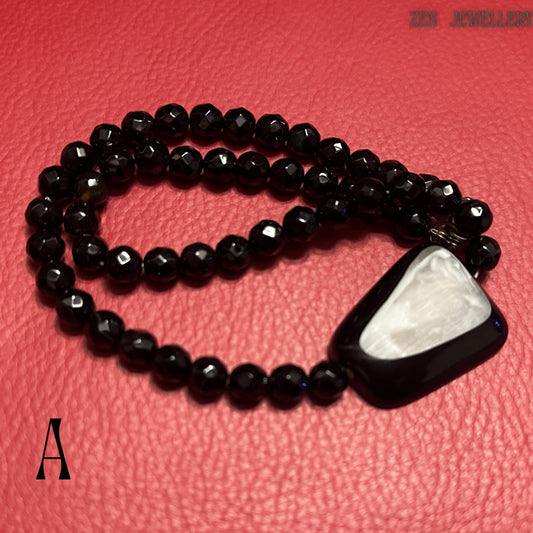Elegant black crystal statement necklace featuring faceted black onyx-style beads arranged symmetrically with a large asymmetrical geometric pendant positioned at the front. The pendant combines a glossy black outer frame with a luminous white shell-like centrepiece that catches light softly. The matching bracelet complements the necklace with the same polished faceted bead design, creating a coordinated luxury jewellery set perfect for formal ou