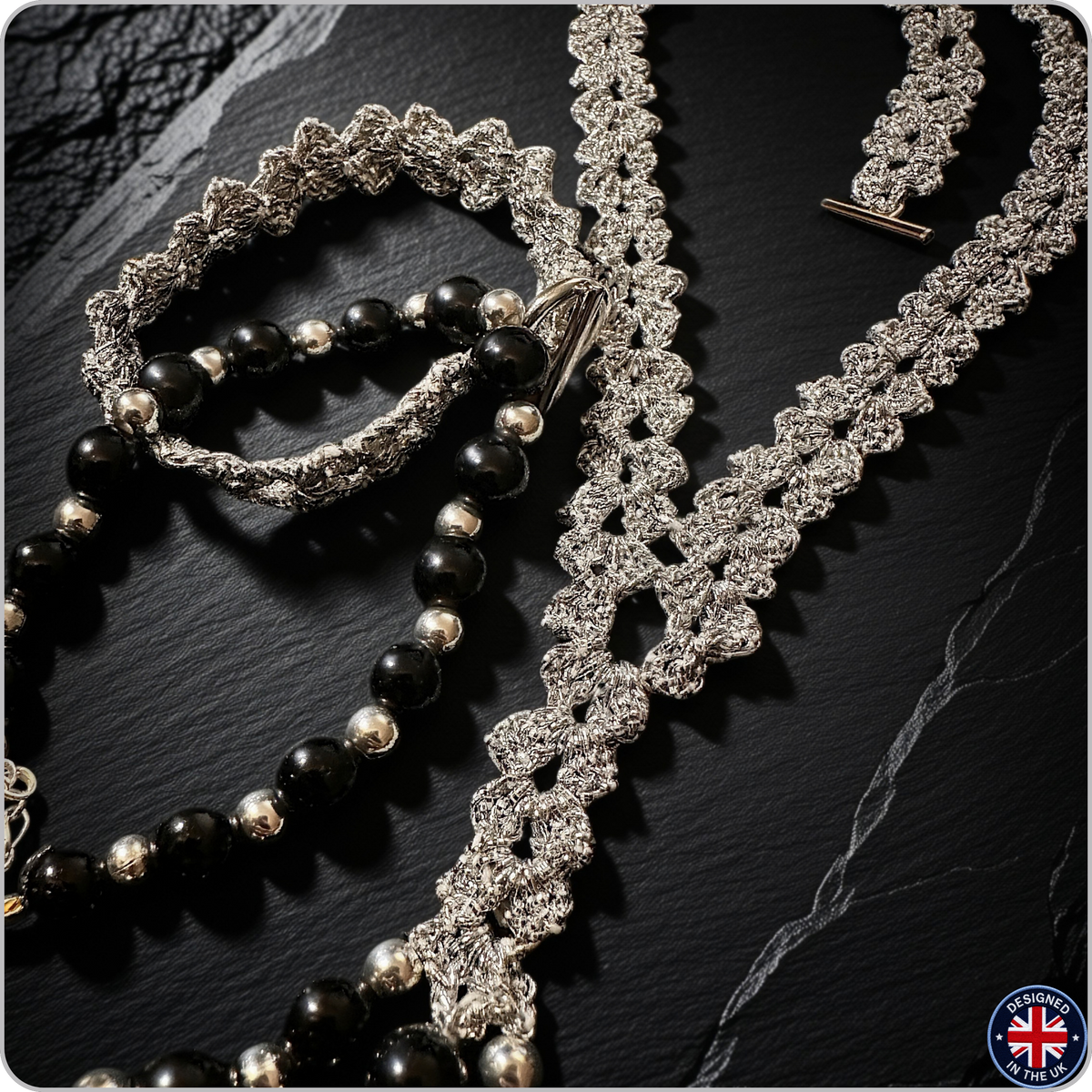 A close-up product shot of jewelry laid on a dark stone surface: a long silver-toned beaded necklace with a toggle clasp, a matching silver-toned bracelet, and a beaded bracelet with glossy black round beads and small metallic silver accents. No person or real environment is present, so this is Product/Packaging rather than Lifestyle.