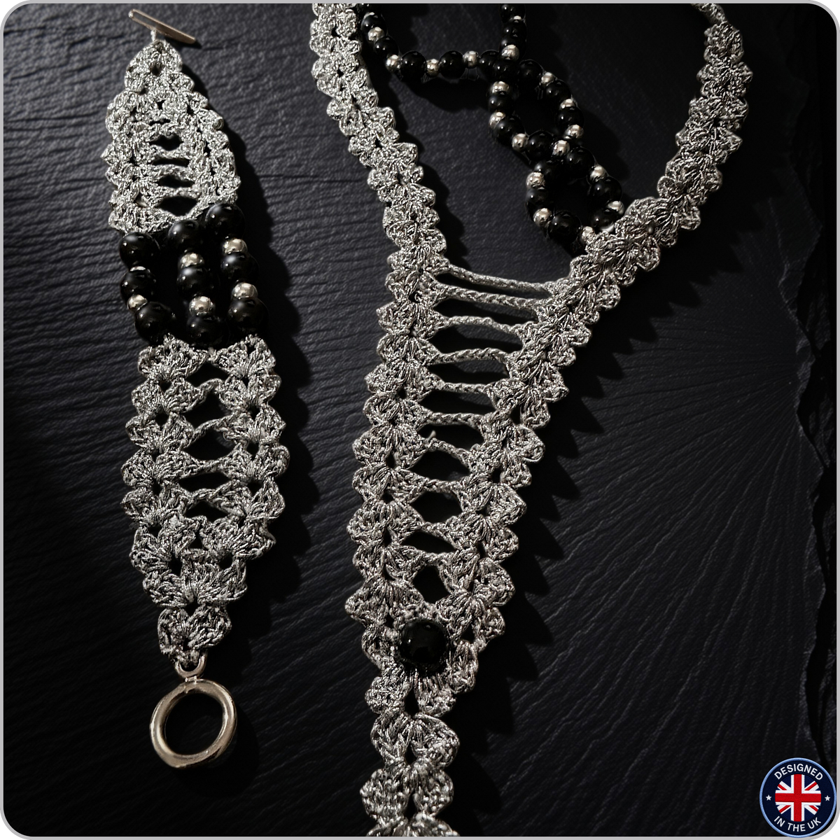 A handmade silver-toned crochet necklace and matching drop earrings with black bead accents, displayed on a dark slate background.