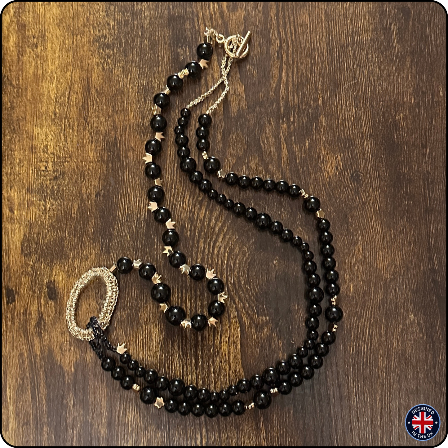A long beaded necklace with black faceted beads and small gold star accents, laid out on a dark brown wooden surface. The necklace has a gold-toned clasp and a matching gold-toned ring detail near one end.