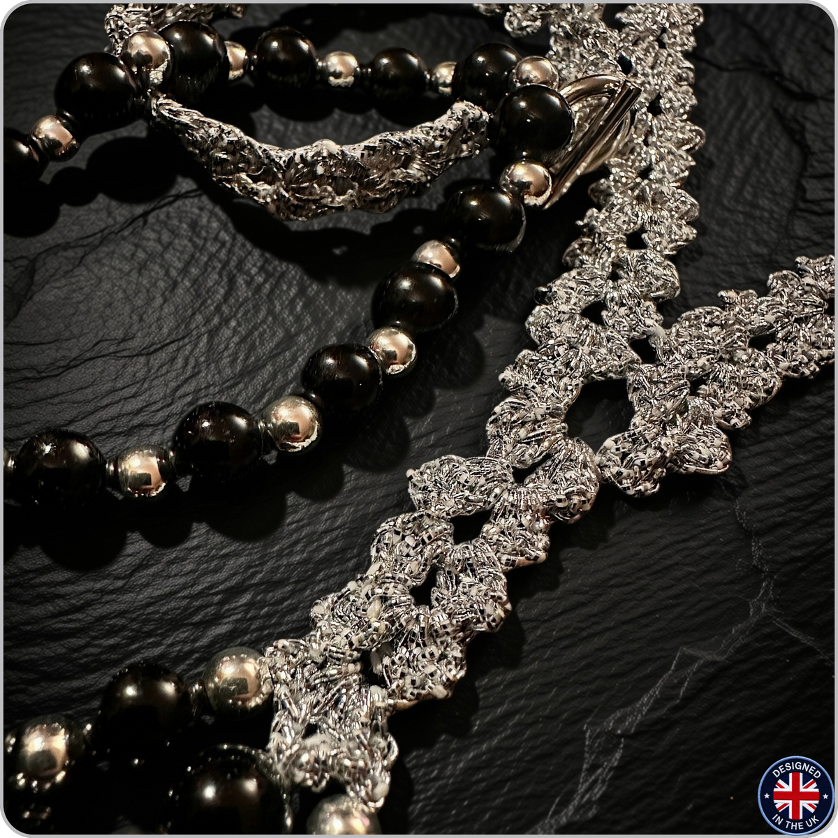 A close-up of a beaded necklace with glossy black oval beads, polished silver spacer beads, and a chunky silver textured chain, displayed on a dark textured surface.