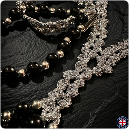 A close-up of a beaded necklace with glossy black oval beads, polished silver spacer beads, and a chunky silver textured chain, displayed on a dark textured surface.