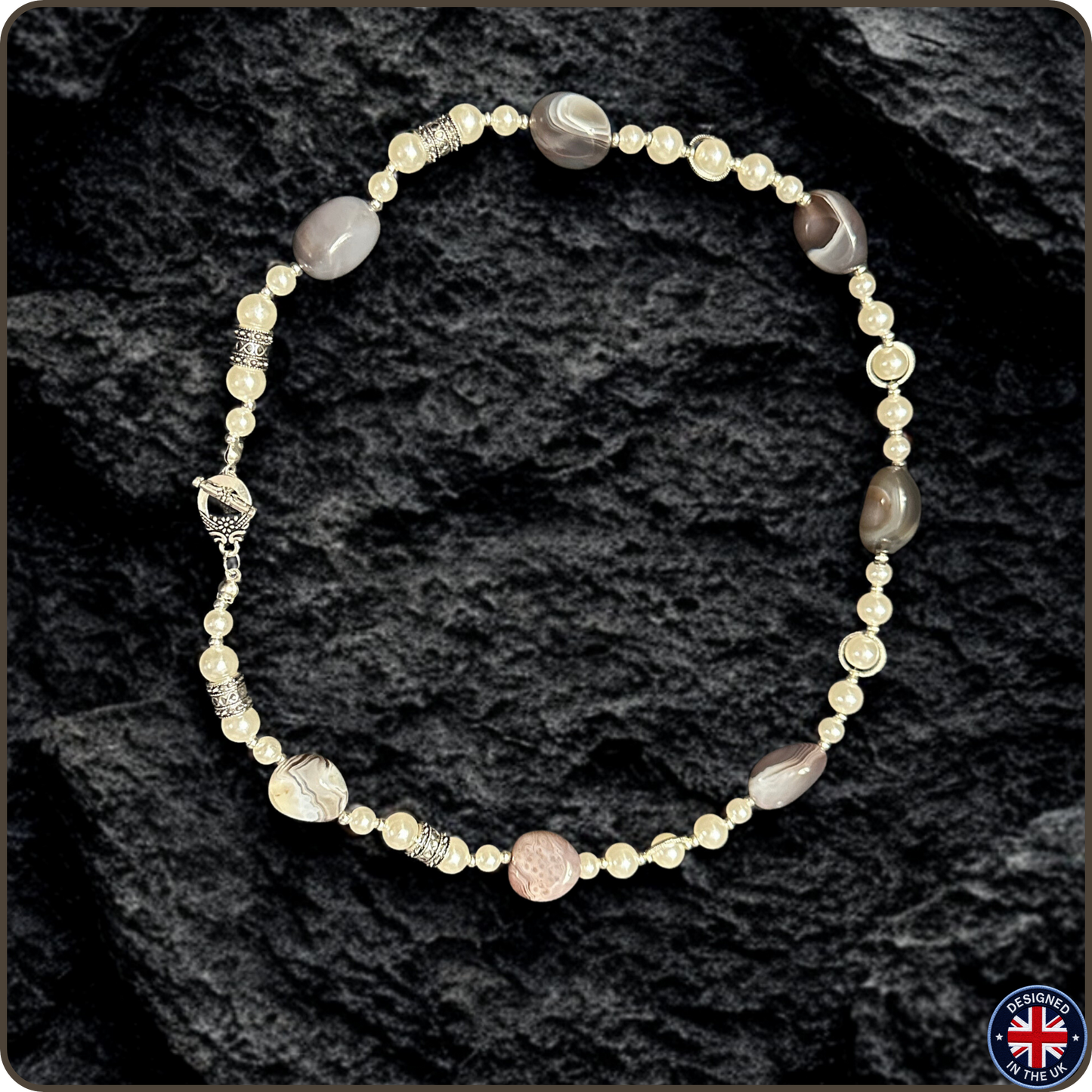 A beaded necklace with a mix of cream, gray, and brown polished beads, silver spacer beads, and a small pinkish-beige round bead, displayed on a dark textured rock background.