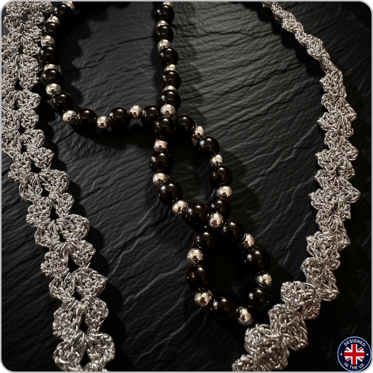 A close-up of a beaded necklace with glossy black and polished silver beads, styled with two silver lace trim strands on a dark textured background.