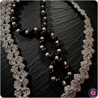 A close-up of a beaded necklace with glossy black and polished silver beads, styled with two silver lace trim strands on a dark textured background.