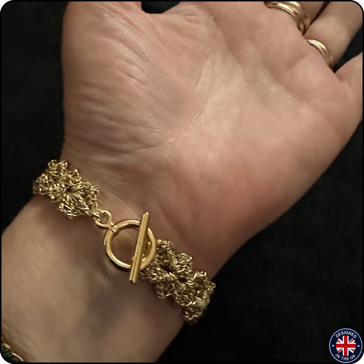 A gold-toned textured bracelet with a toggle clasp worn on a person's wrist, shown against a dark background. The person's hand and part of the forearm are visible.