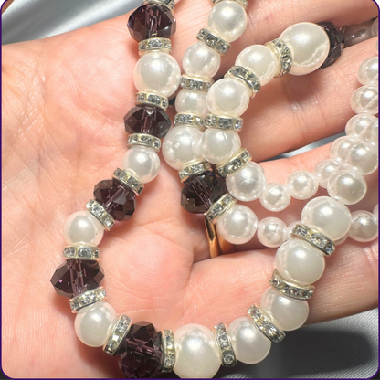 A close-up of a beaded necklace draped across a hand. The necklace features large white faux-pearl round beads, faceted dark plum crystal beads, and small silver-toned spacers set with clear rhinestones.