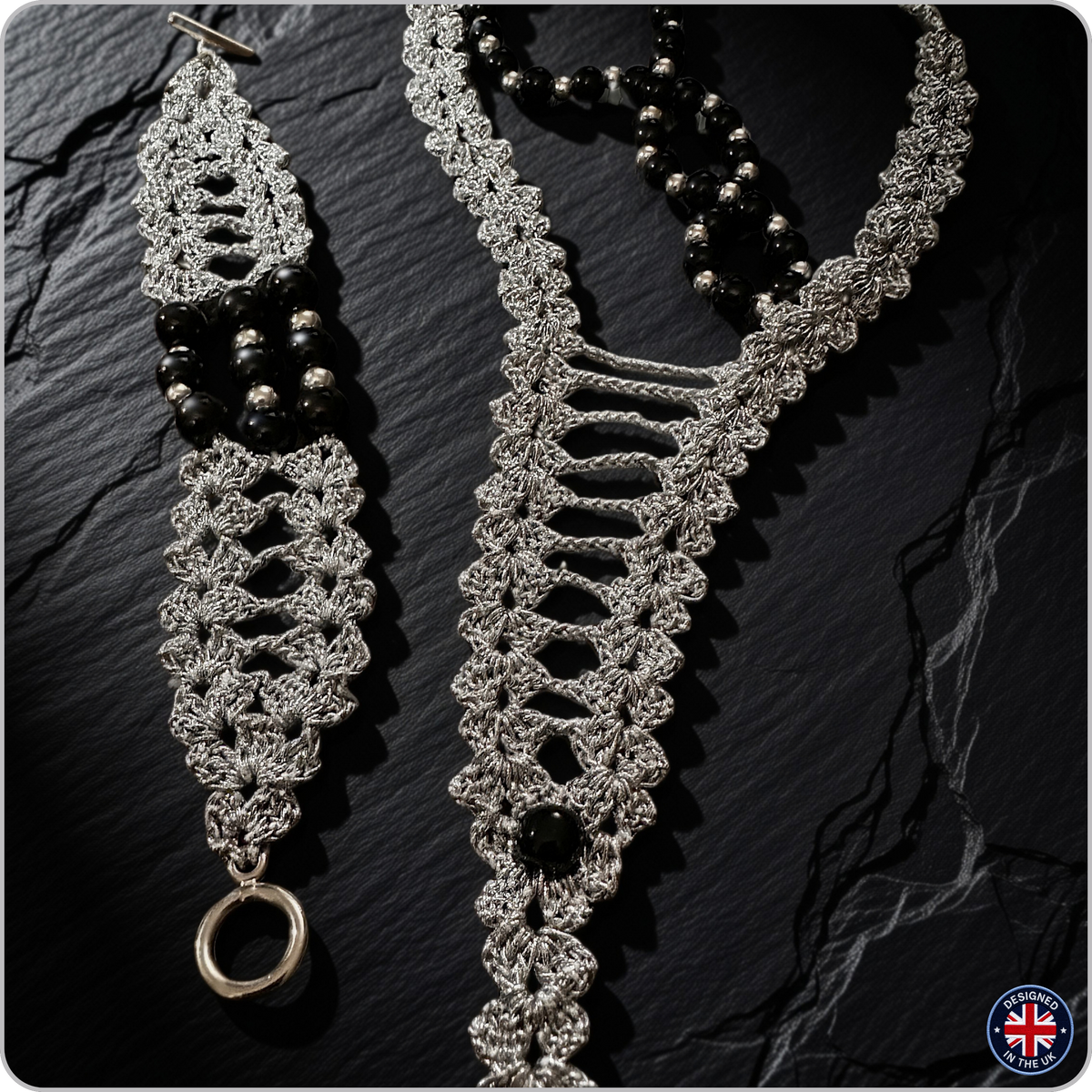 A silver-toned crochet or macrame jewelry set displayed on a dark textured stone surface, including a long necklace and a matching bracelet or anklet with black beads and a ring detail.