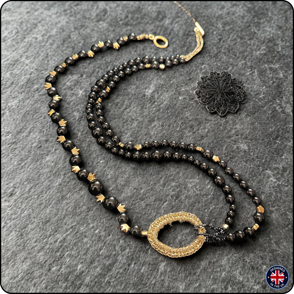 A double-strand beaded necklace with black faceted beads, small gold spacer beads, and a central gold-toned textured oval pendant, displayed on a dark gray textured background next to a black lace charm.
