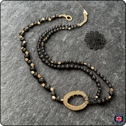 A double-strand beaded necklace with black faceted beads, small gold spacer beads, and a central gold-toned textured oval pendant, displayed on a dark gray textured background next to a black lace charm.