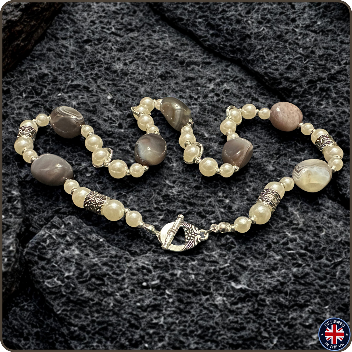 A beaded bracelet and matching ring featuring cream-colored faux pearls, gray agate beads, and textured silver-toned spacer beads, displayed on a dark textured stone background.
