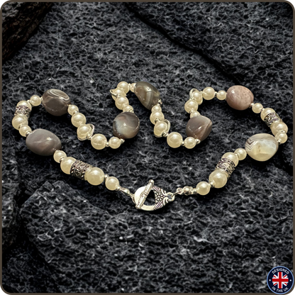 A beaded bracelet and matching ring featuring cream-colored faux pearls, gray agate beads, and textured silver-toned spacer beads, displayed on a dark textured stone background.
