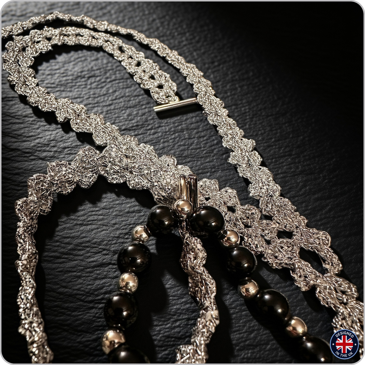 A silver-toned beaded necklace with intricate lace-like woven details and glossy black beads, displayed on a dark textured surface.
