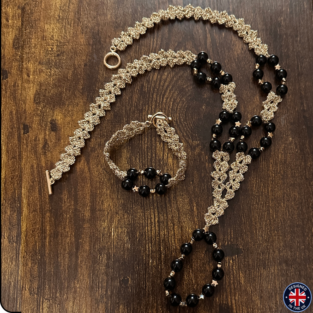 A jewelry set featuring a long beaded necklace, a matching bracelet, and a ring, styled on a dark wood background. The jewelry has a gold-toned floral lace chain with black faceted beads and small gold star accents.