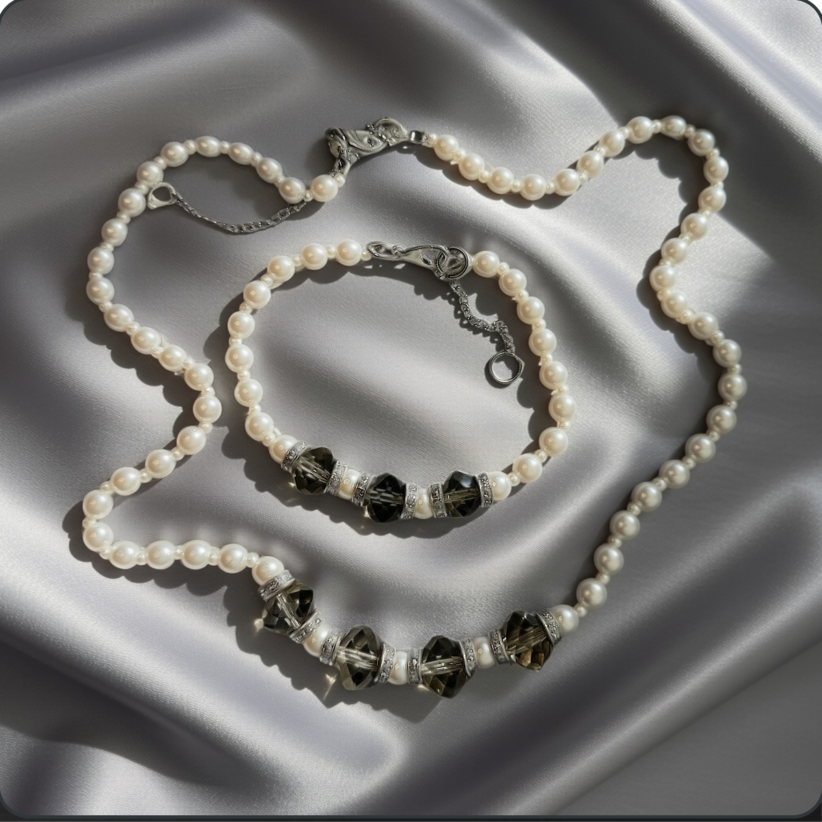 A pearl-bead necklace and matching bracelet with dark faceted crystal beads and silver-toned accents, arranged on draped satin fabric.