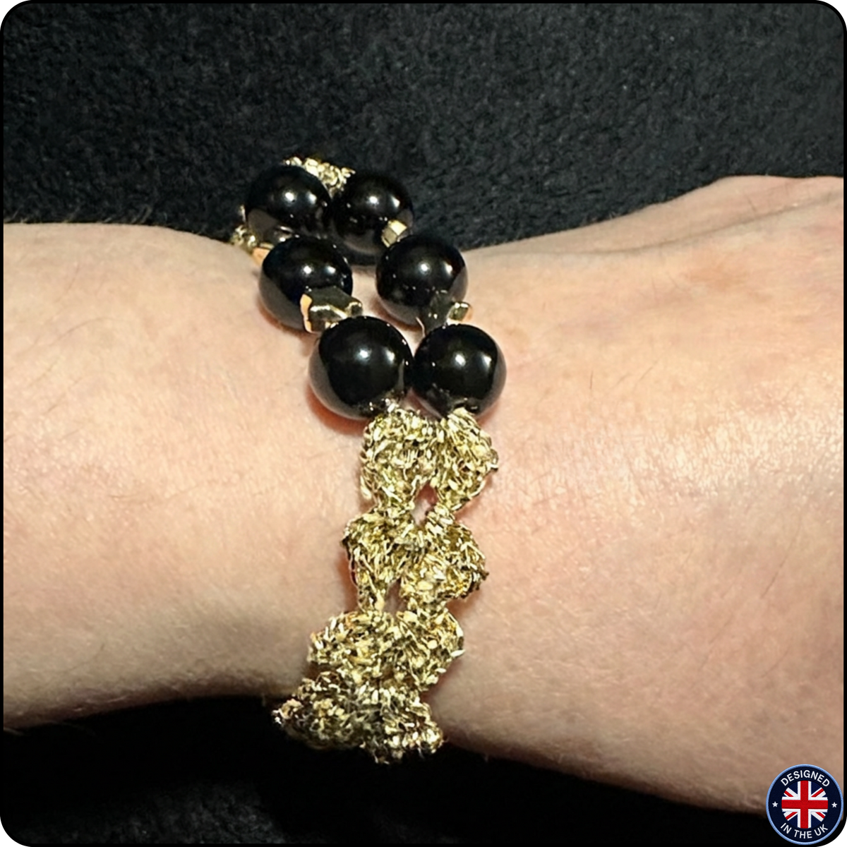 A close-up of a person's wrist wearing a beaded bracelet with glossy black round beads and a chunky textured gold woven section, set against a dark background.