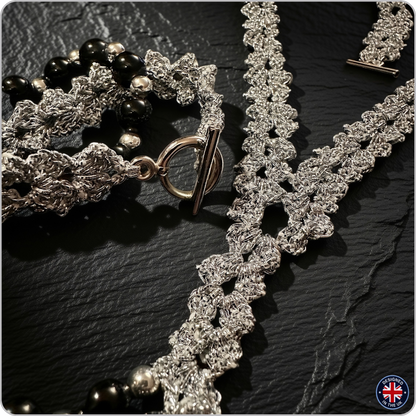 A close-up of jewelry laid on a dark textured surface, including a silver-toned beaded necklace with black beads, a silver-toned beaded bracelet with black beads, and a silver-toned textured chain necklace with a toggle clasp.