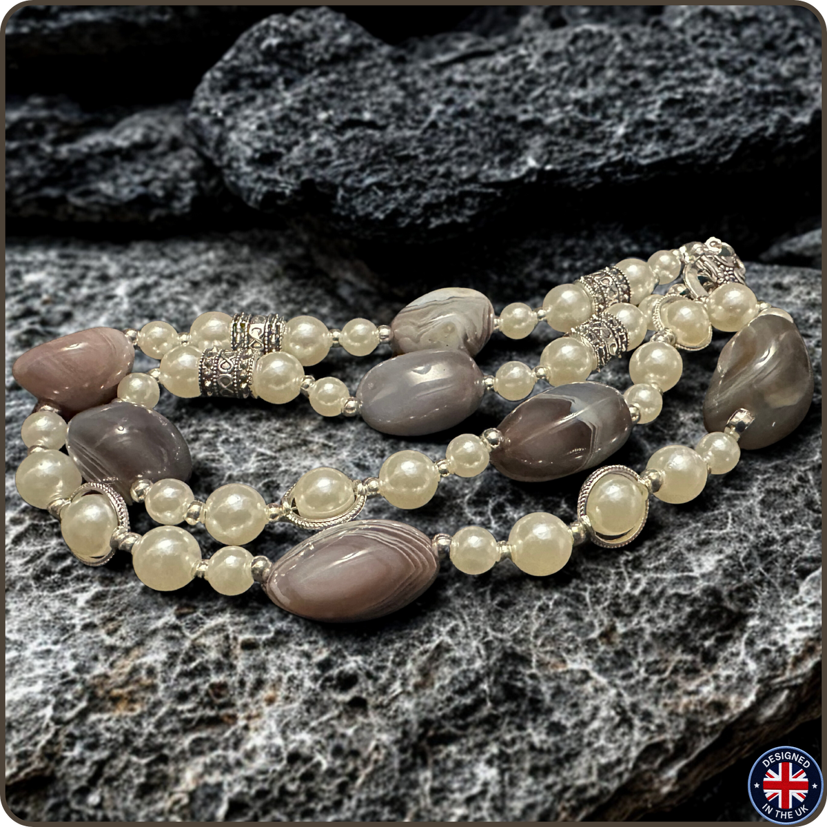 A beaded necklace with oval agate-like stones in gray and taupe, cream pearl-like beads, and silver-toned spacer details, arranged on dark gray rocks.