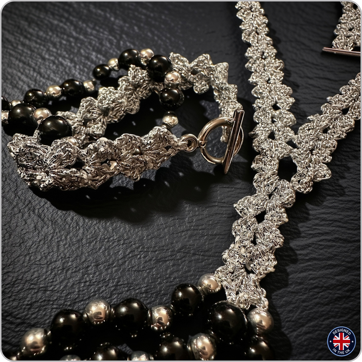 A close-up of a jewelry set featuring a silver-toned floral lace necklace with a toggle clasp, a matching silver-toned bracelet, and strands of black and silver beads, all arranged on a dark textured surface.
