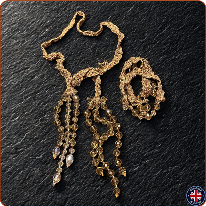 A gold-toned beaded and textured accessory set laid on a dark textured background, featuring a long dangling piece with multiple strands of faceted beads and a smaller matching coiled piece. 