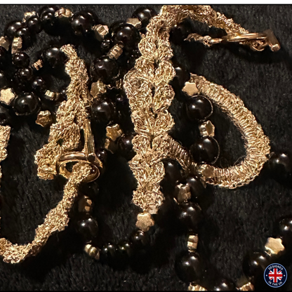 A close-up of a vintage-style beaded necklace featuring glossy black round beads, textured gold chain links, and small metallic gold star-shaped beads, laid out on a dark textured background.