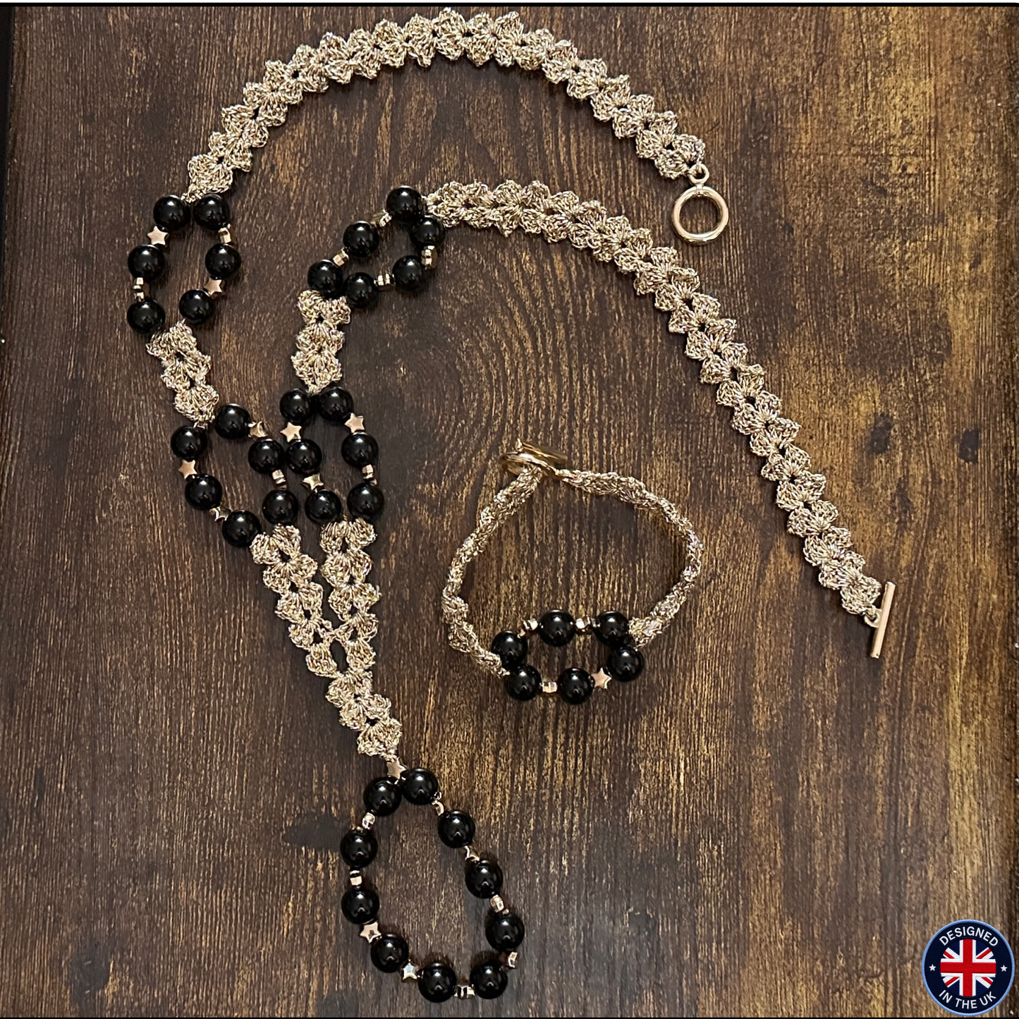A jewelry set laid flat on a dark wood surface, including a long beaded necklace, a matching bracelet, and a smaller beaded bracelet or anklet. The pieces feature black faceted beads and metallic gold-toned woven chain details with small star accents.