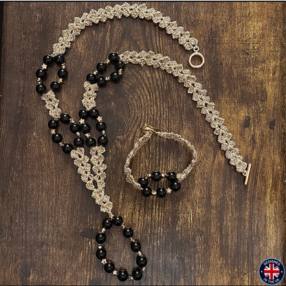 A jewelry set laid flat on a dark wood surface, including a long beaded necklace, a matching bracelet, and a smaller beaded bracelet or anklet. The pieces feature black faceted beads and metallic gold-toned woven chain details with small star accents.
