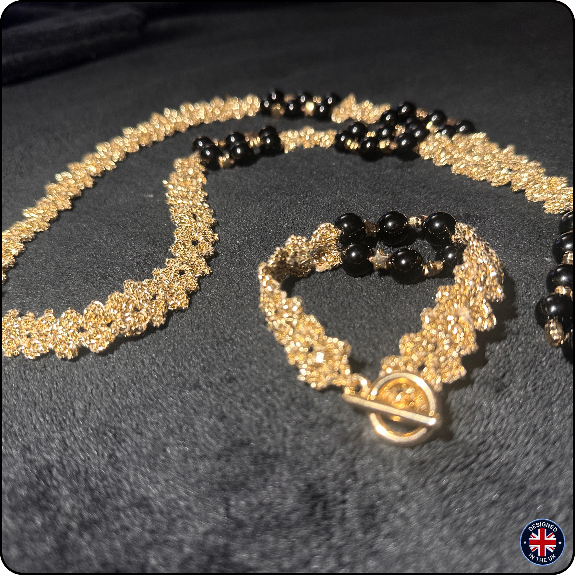 A gold and black beaded jewelry set featuring a textured floral-patterned necklace and matching bracelet with black beads, displayed on a dark textured fabric background.