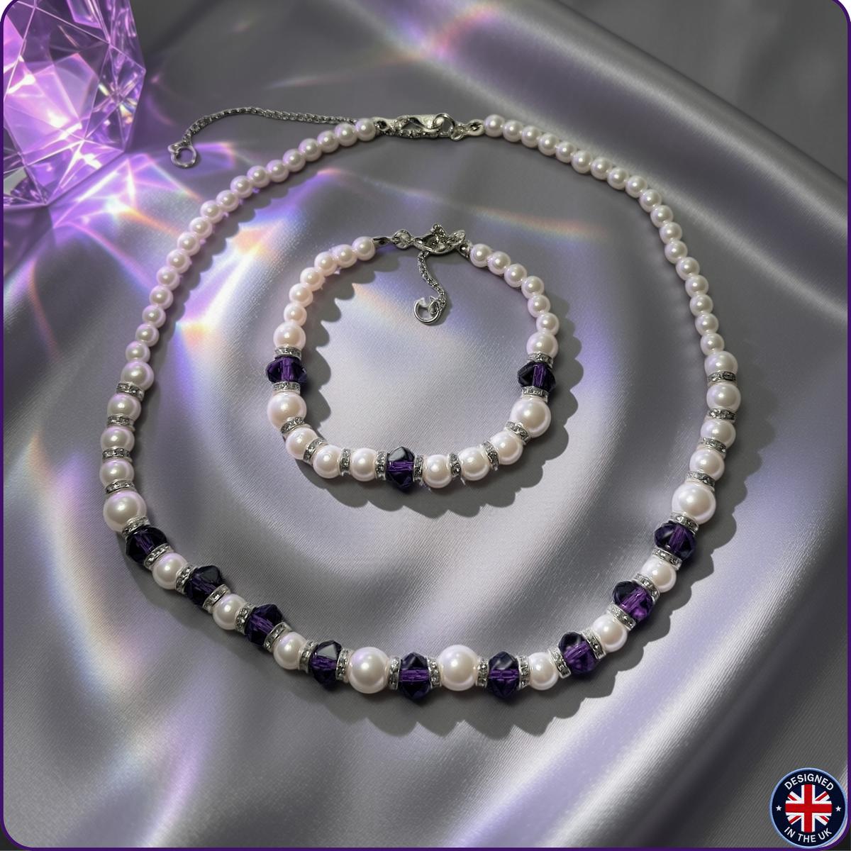 A pearl necklace and matching pearl bracelet set with silver-toned chain links and faceted purple crystal beads, arranged on a shiny silver satin fabric background with a purple gem prop in the corner.