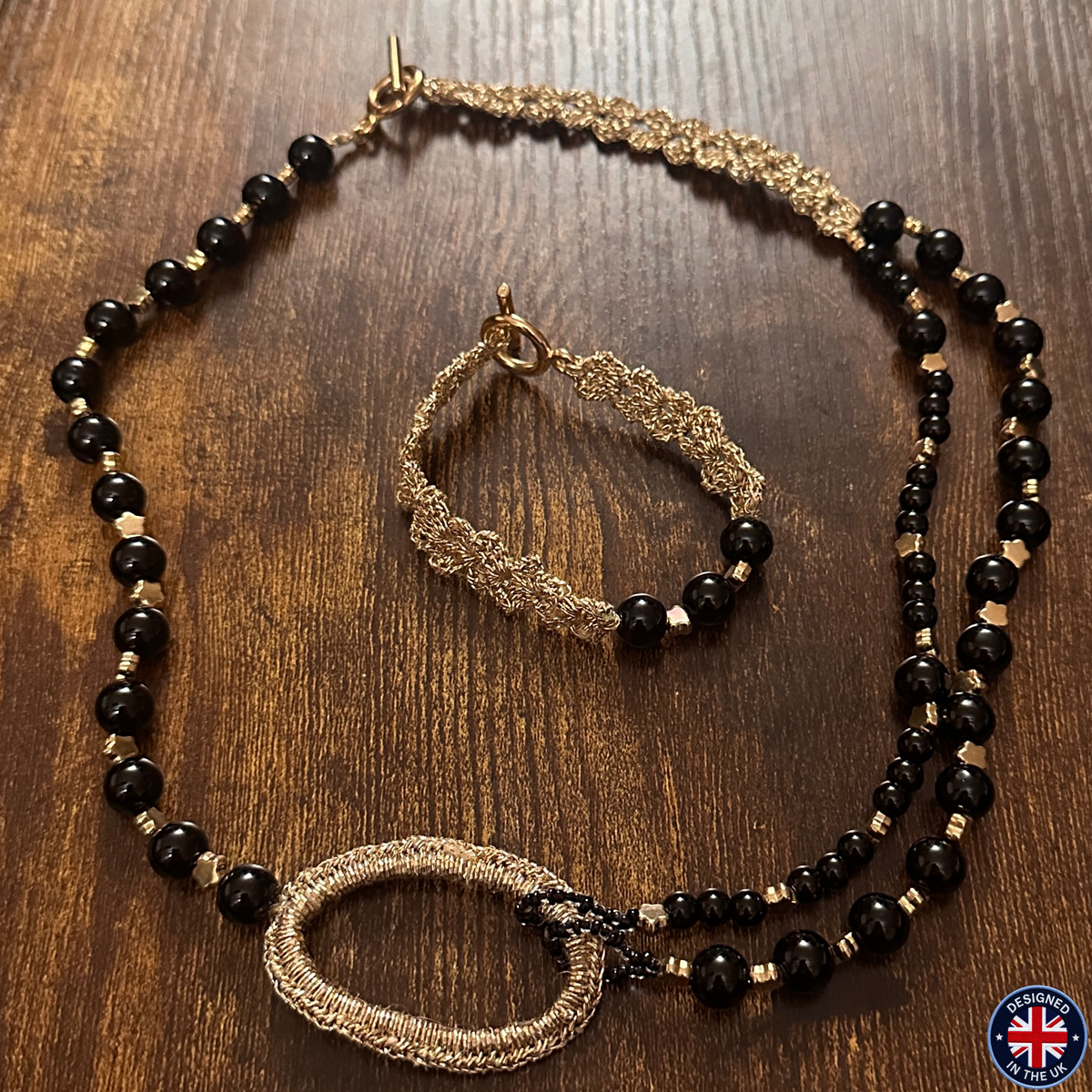 A beaded necklace and matching bracelet set displayed on a dark wood background. The jewelry features black faceted beads, gold-toned chain sections, and small metallic gold spacer beads.
