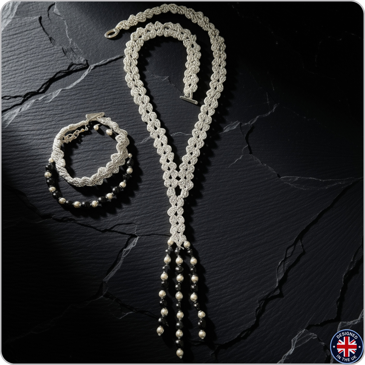 A handmade macrame necklace and matching bracelet set displayed on a dark textured slate background. The macrame is cream-colored, while the beaded strands at the bottom feature cream beads and dark gray beads.