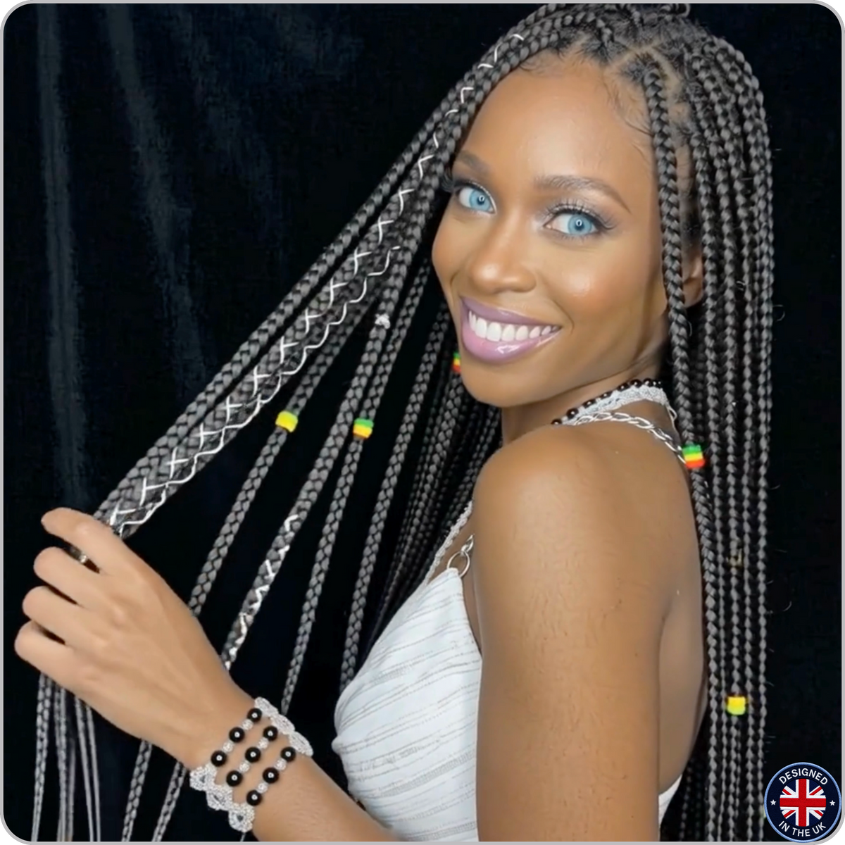 A woman with long black box braids decorated with silver braided accents and small colorful beads, wearing a white sleeveless top against a black curtain background.