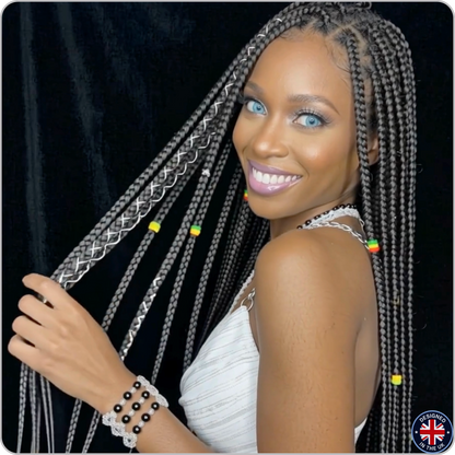 A woman with long black box braids decorated with silver braided accents and small colorful beads, wearing a white sleeveless top against a black curtain background.