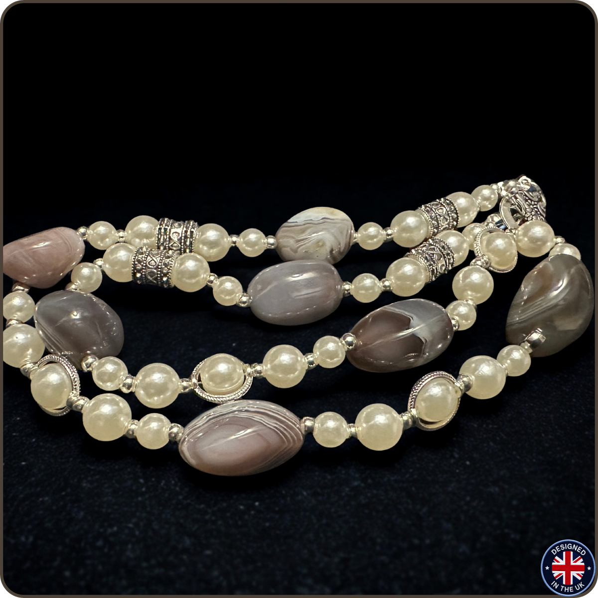 A beaded necklace featuring cream-colored faux pearls, polished oval agate stones in shades of gray, brown, and white, and ornate silver-toned spacer beads, displayed on a black textured background.