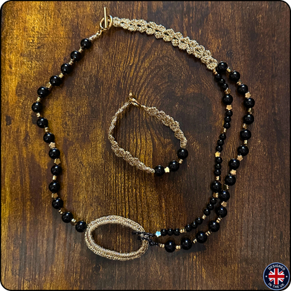 A jewelry set featuring a beaded necklace and matching bracelet laid out on a dark wood background. The jewelry has black faceted beads, gold-toned woven sections, and small gold spacer beads.