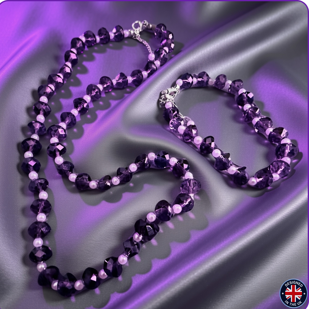 A purple beaded necklace and matching bracelet displayed on draped purple satin fabric. The jewelry features faceted dark purple beads, light lavender beads, and small white pearl-like accents, with silver-tone clasps.
