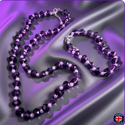 A purple beaded necklace and matching bracelet displayed on draped purple satin fabric. The jewelry features faceted dark purple beads, light lavender beads, and small white pearl-like accents, with silver-tone clasps.