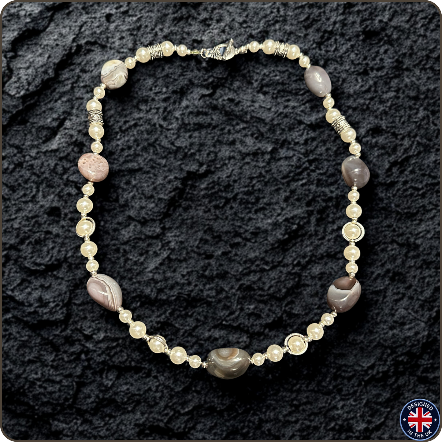 A beaded necklace featuring cream-colored faux pearls, silver-toned spacer beads, and polished stone beads in gray, brown, and pinkish tones, displayed on a dark textured rock background.