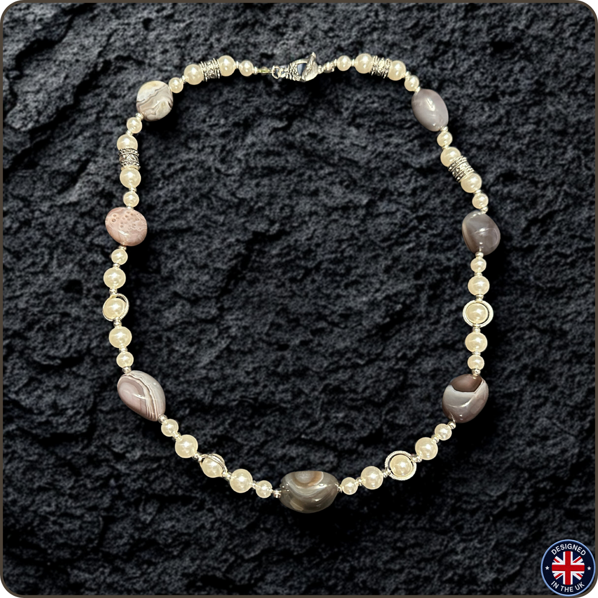 A beaded necklace featuring cream-colored faux pearls, silver-toned spacer beads, and polished stone beads in gray, brown, and pinkish tones, displayed on a dark textured rock background.