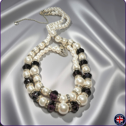 A pearl statement necklace with faceted white and purple crystal beads, silver spacer rings,  displayed on a draped silver satin fabric.