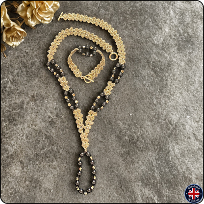 A gold and black beaded necklace set with a matching bracelet, displayed on a textured gray background with decorative gold flowers in the corner.