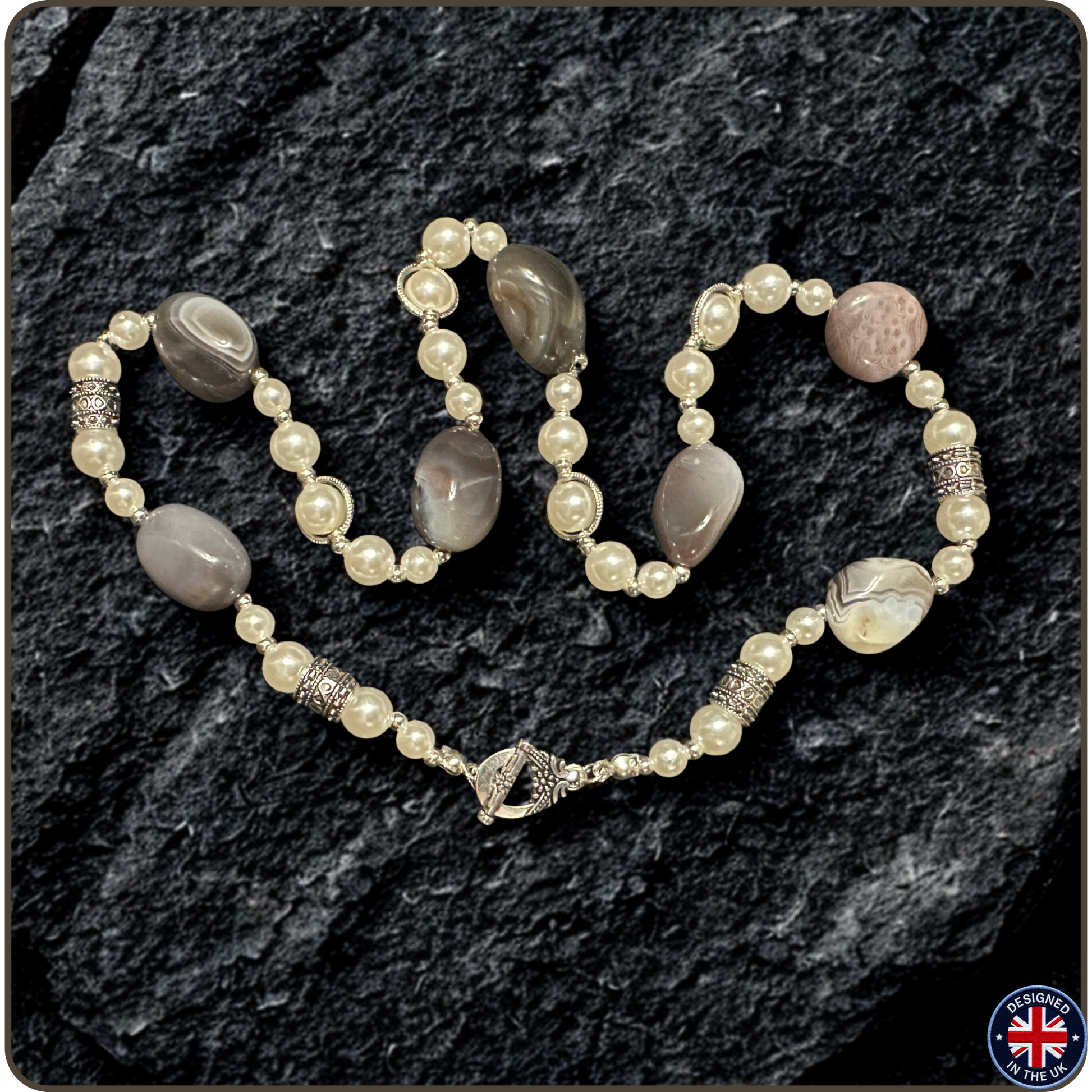A beaded necklace with cream-colored faux pearls, gray agate-like oval beads, and a pinkish agate bead, accented with silver-toned spacer beads, displayed on a dark textured rock background.