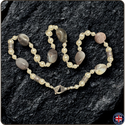 A beaded necklace with cream-colored faux pearls, gray agate-like oval beads, and a pinkish agate bead, accented with silver-toned spacer beads, displayed on a dark textured rock background.