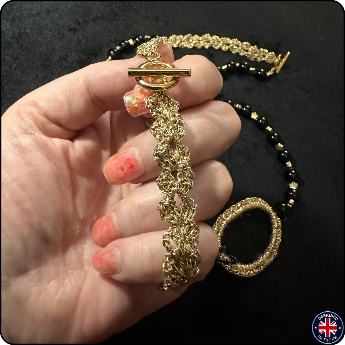 A close-up of a hand holding a gold-toned beaded bracelet with a toggle clasp, with a matching beaded necklace draped in the background on a black surface.