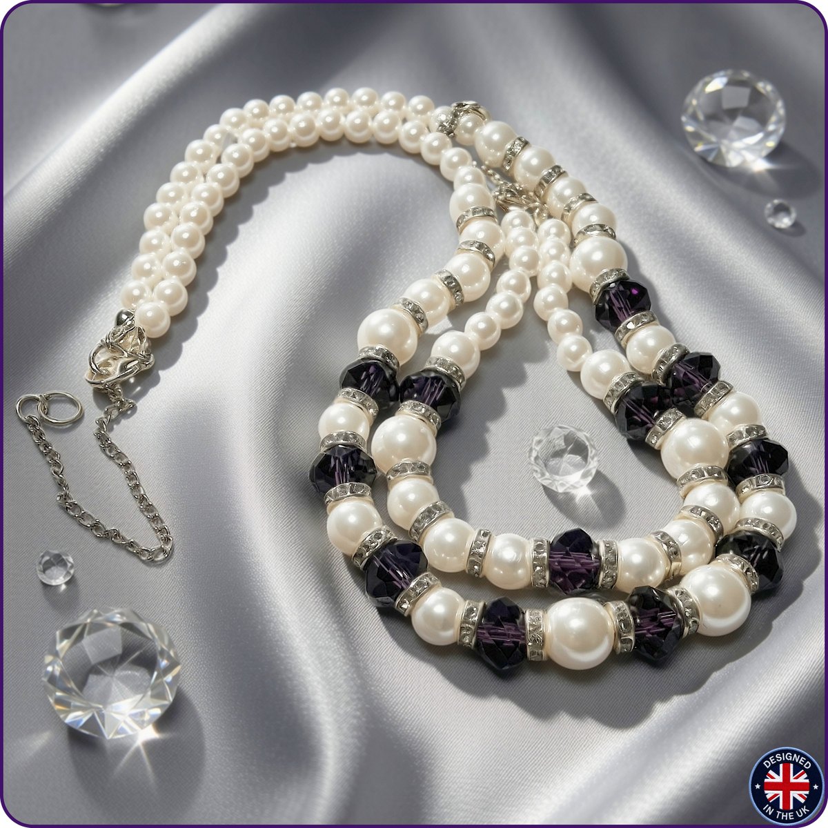 A pearl necklace with purple faceted crystal beads and silver-toned rhinestone spacers, displayed on a satin fabric background with decorative clear crystal props.