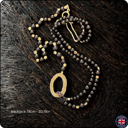 A beaded necklace with dark gray faceted beads, gold star accents, and a large oval pendant covered in small clear stones, displayed on a dark wood background. Text at the bottom reads 'Necklace 78cm - 30.