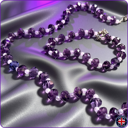 A beaded necklace and matching bracelet made of faceted purple crystal beads separated by small white beads, displayed on draped satin fabric with purple lighting. A silver-toned clasp is visible on the bracelet.