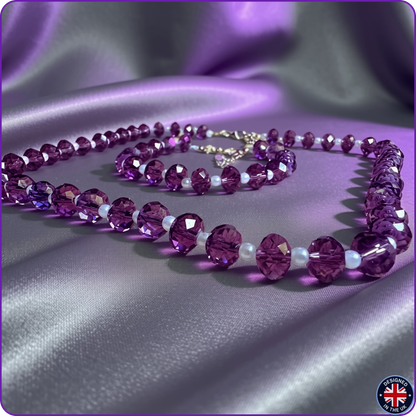 A beaded necklace and matching bracelet made of faceted purple crystal beads separated by small white pearl-like beads, displayed on draped satin fabric. A silver-tone clasp is visible at the back of the necklace.