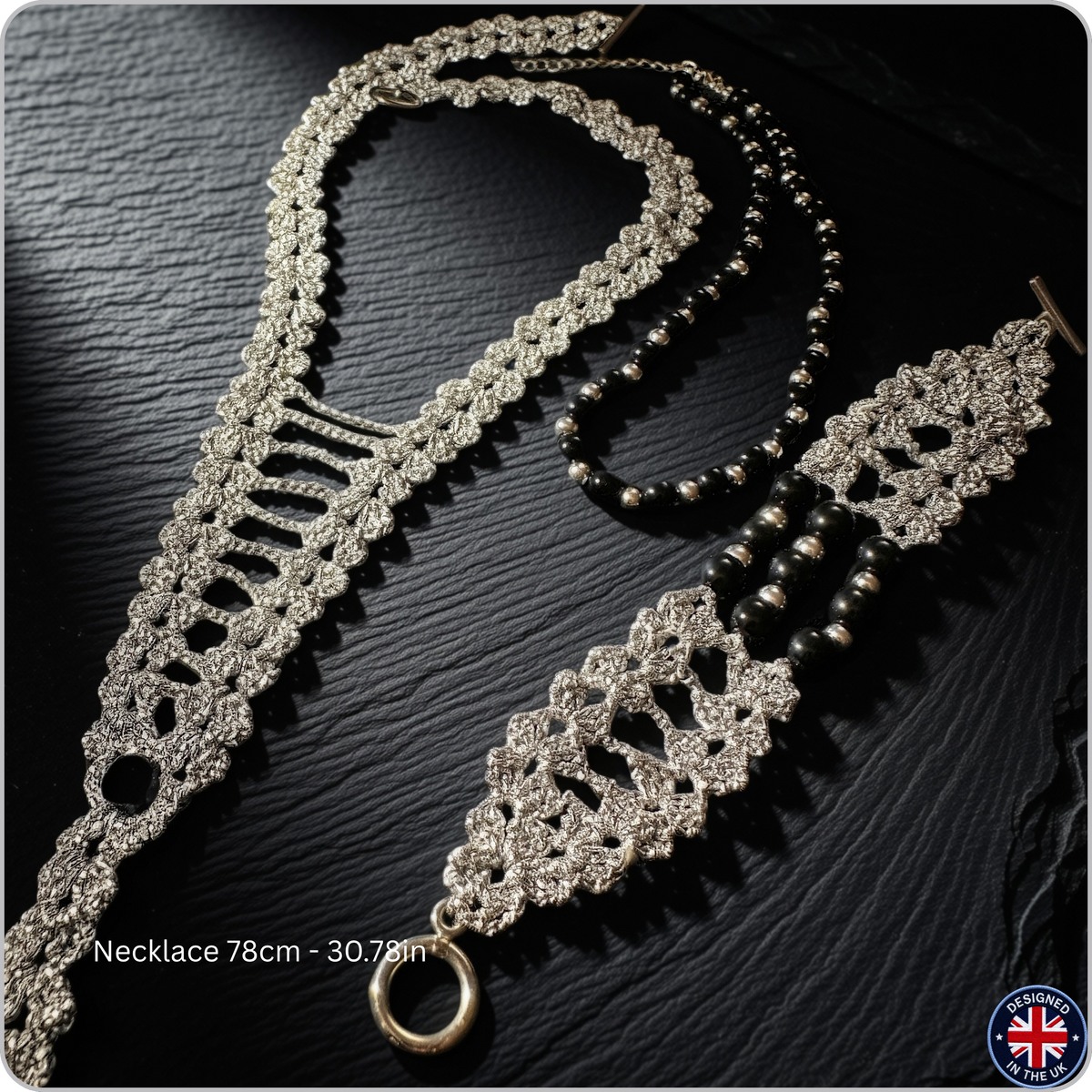 A silver-toned beaded necklace with a matching ornate lace-style pendant and a separate strand of dark beads, arranged on a dark textured surface. White text at the bottom left reads 'Necklace 78cm - 30.